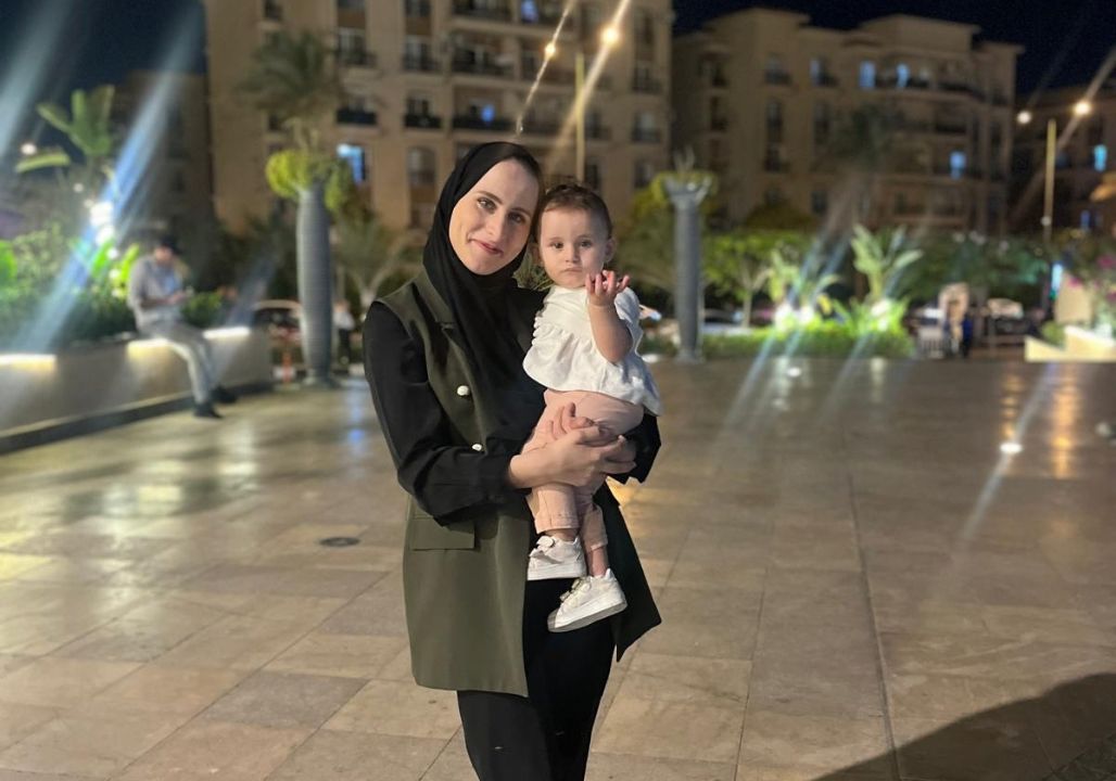 Saja Redwan holding her daughter, Tia, while posing outdoors for a portrait. It’s night, and the streetlamps illuminate a city in the distance.