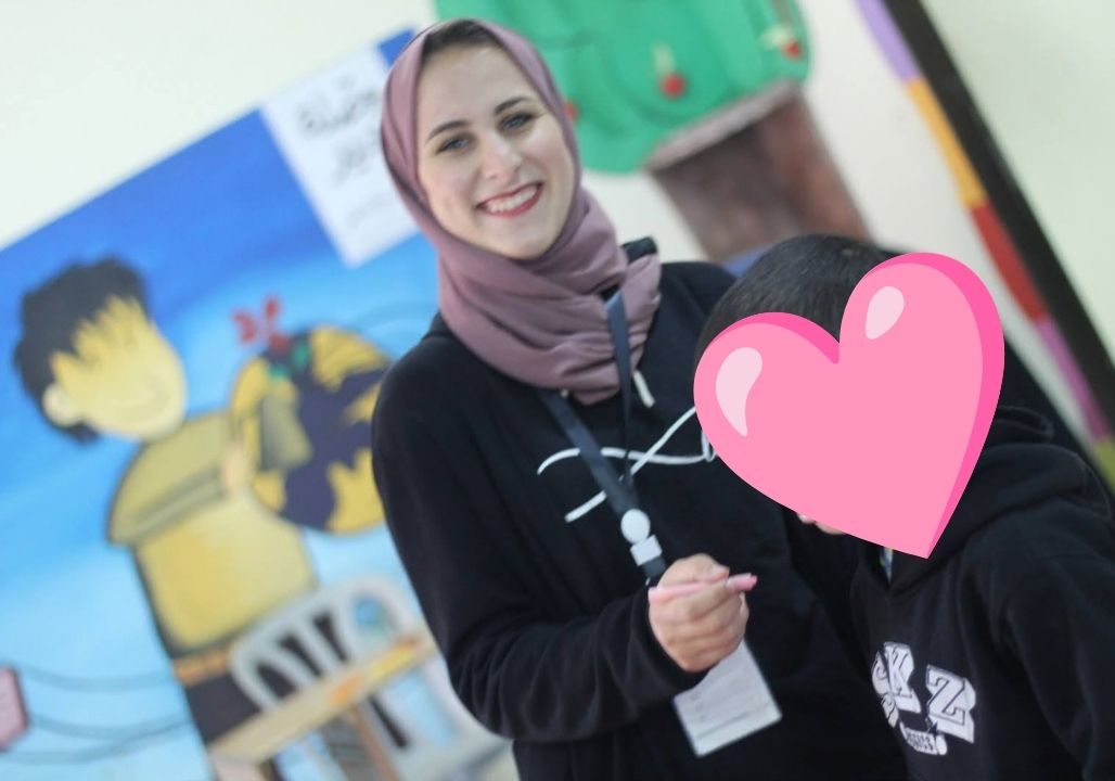 Saja Redwan smiling while standing next to a child she worked with during one of her mentorship programmes. The child’s face is covered with a pink heart emoji to protect their anonymity.