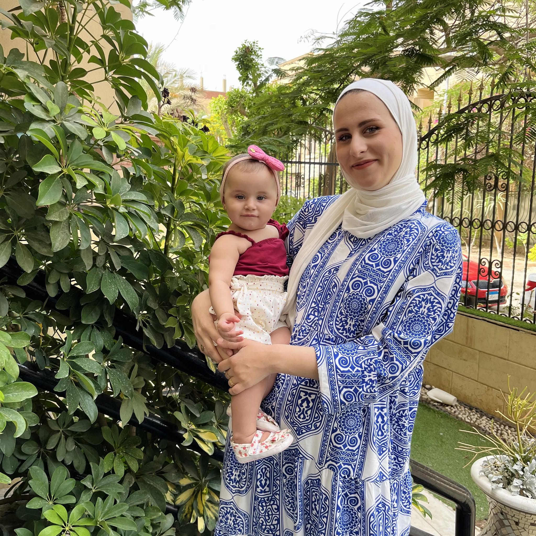 Saja Redwan, freelance writer, marketing professional, and postgraduate student, holding her daughter, Tia, outdoors in front of a leafy plant.